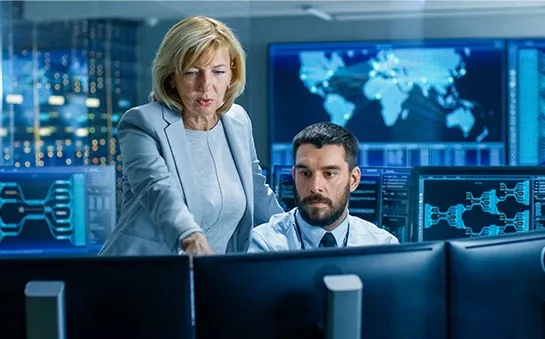 A woman and a man work at multiple computer monitors, engaging in a discussion in a high-tech control room.