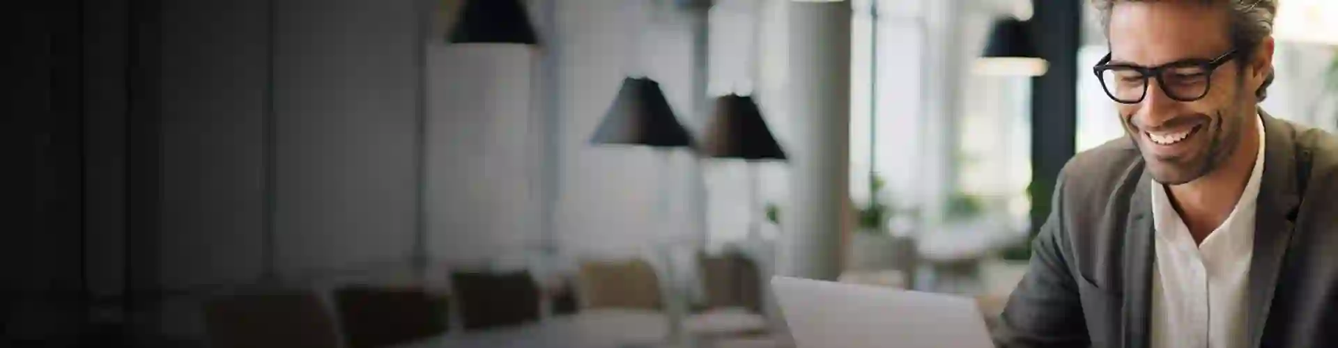A person wearing glasses smiles while looking at a laptop, sitting in a modern, bright office with hanging lamps and plants visible.