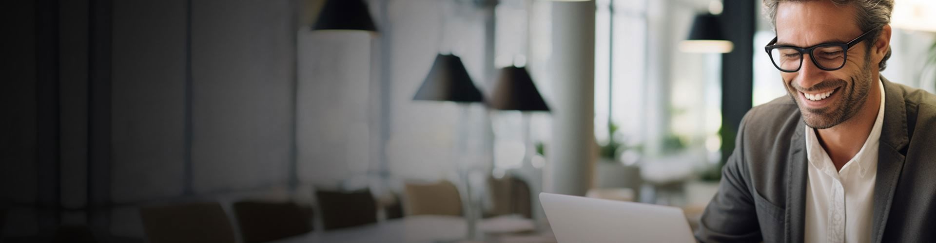 A person wearing glasses smiles while looking at a laptop, sitting in a modern, bright office with hanging lamps and plants visible.